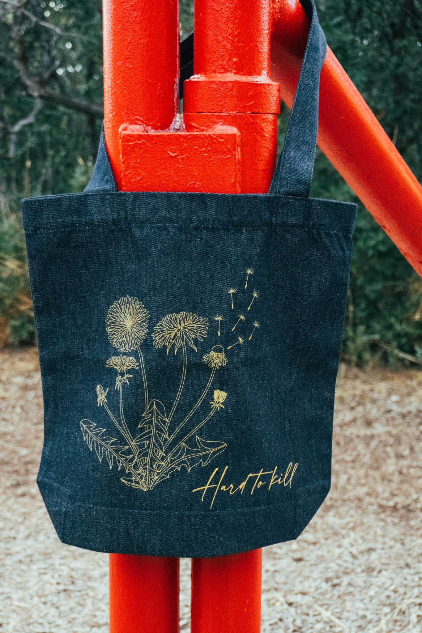 Blue denim tote bag with dandelion design and 'Hard to Kill' text, held by a red gate.