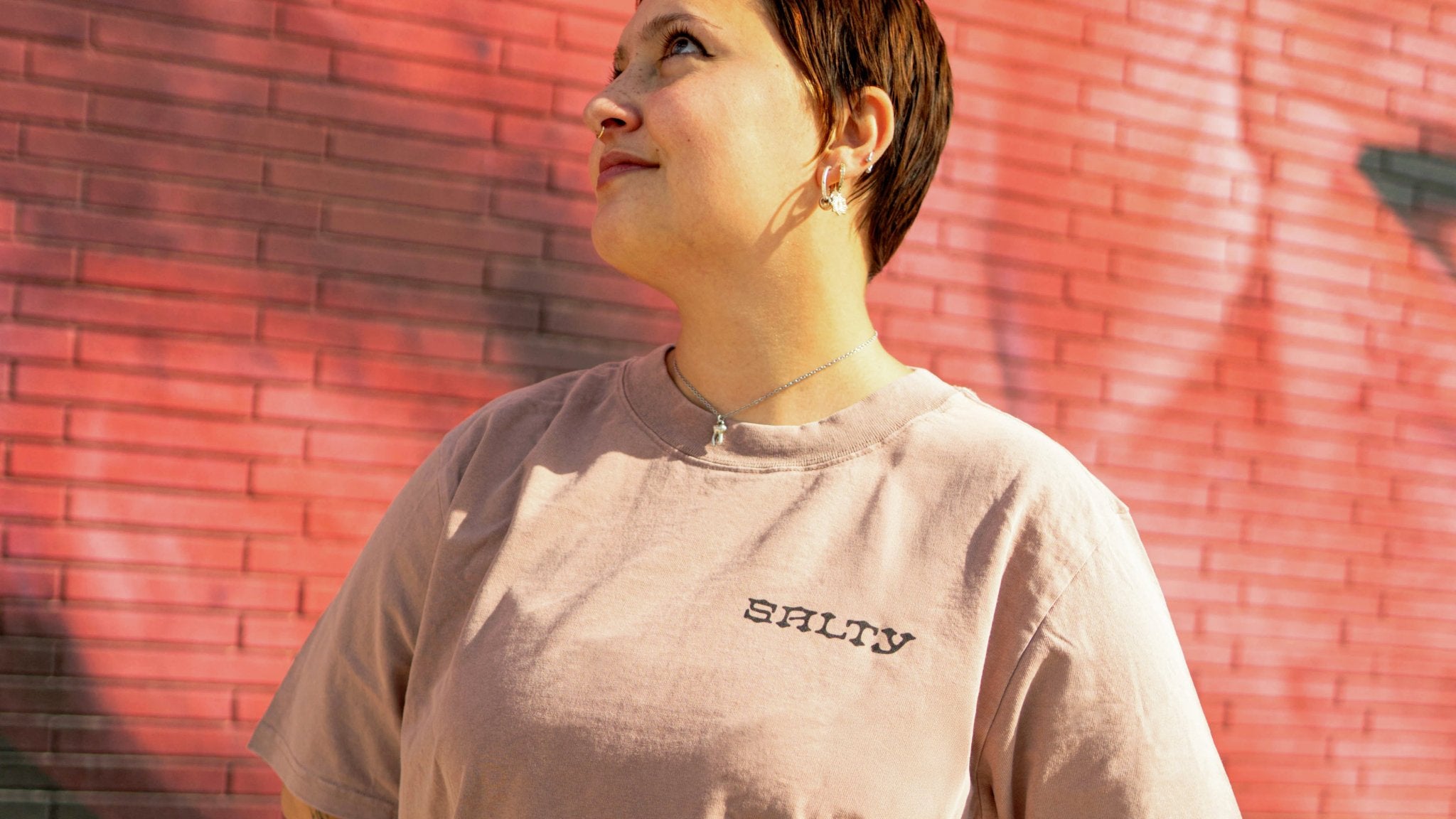 Person wearing a light pink t-shirt with 'SALTY' text against a red brick wall.