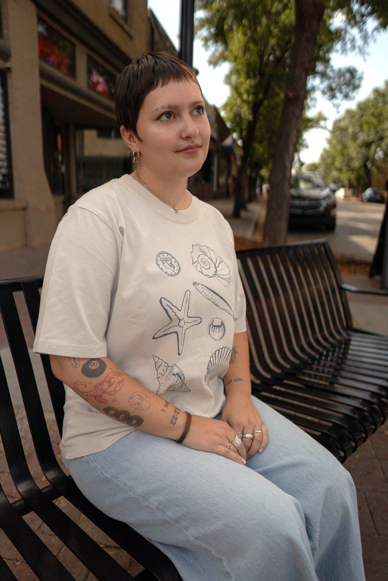 Person sitting on a bench wearing a white t-shirt with graphics of sea shells and light blue jeans.