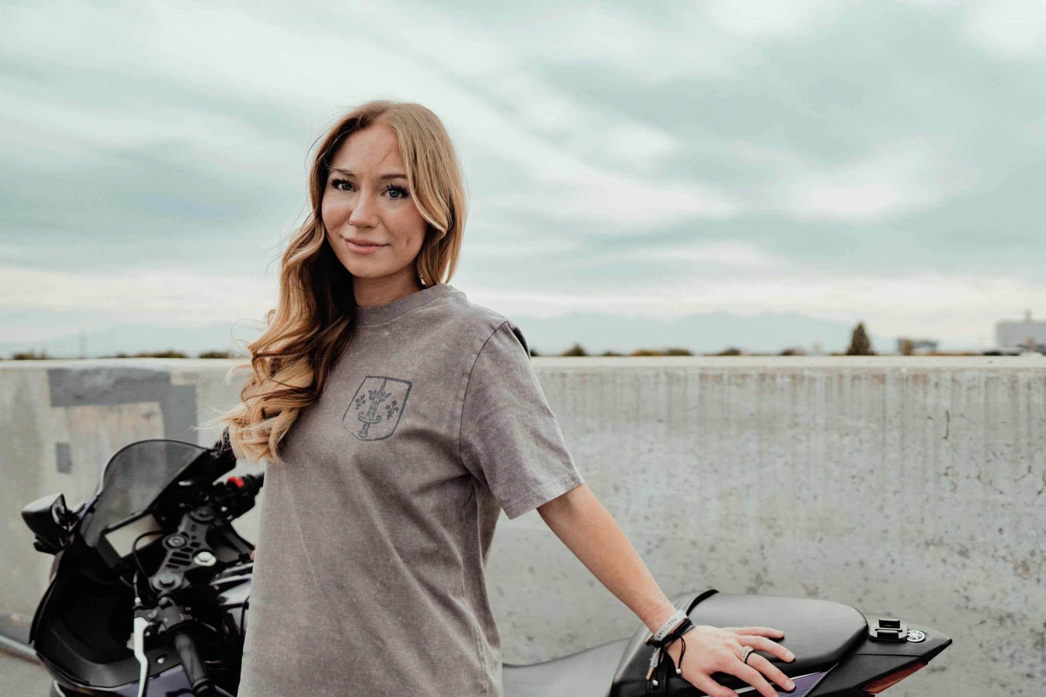 Woman standing next to a motorcycle on a rooftop with a cloudy sky.