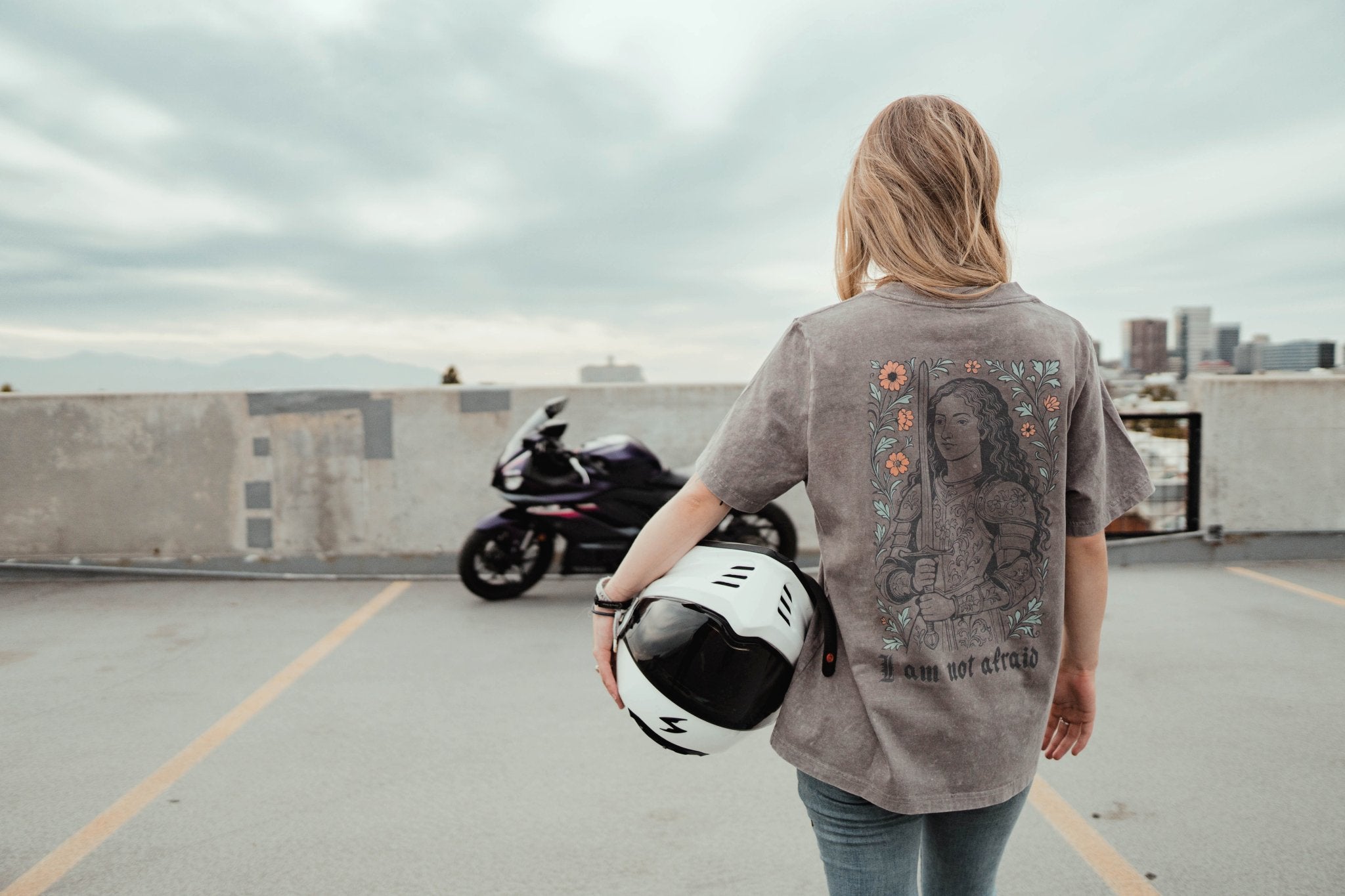 Person holding a helmet on a rooftop with a motorcycle in the background and a shirt that reads 'I am not afraid'