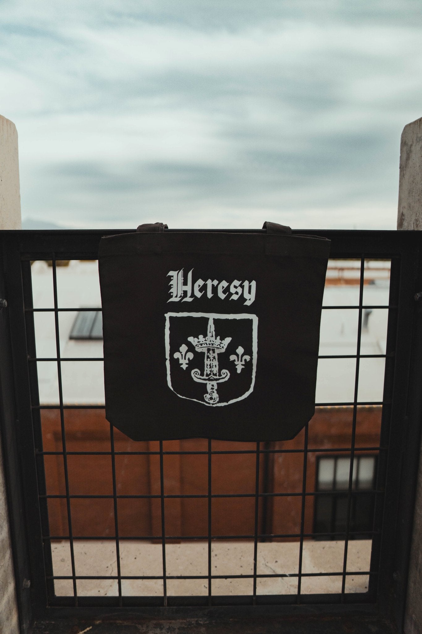 Black tote bag with 'Heresy' text and emblem hanging on a railing with a cloudy sky background.