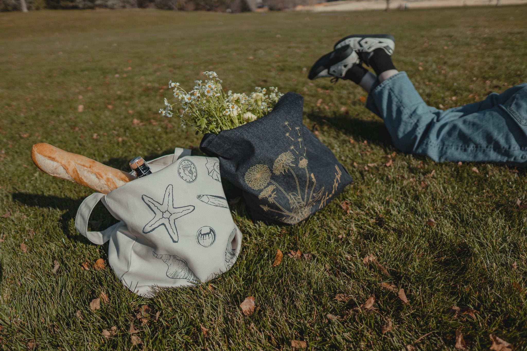 Person lying on grass with a baguette, flowers in a tote bag with nature-themed design.