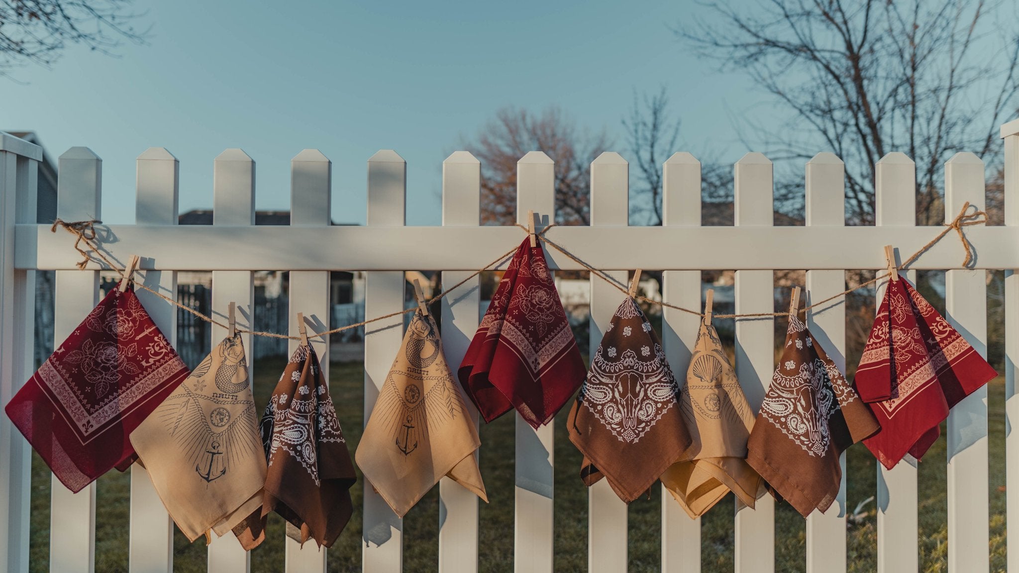 Bandanas hanging on a clothesline against a white picket fence with a blue sky background.