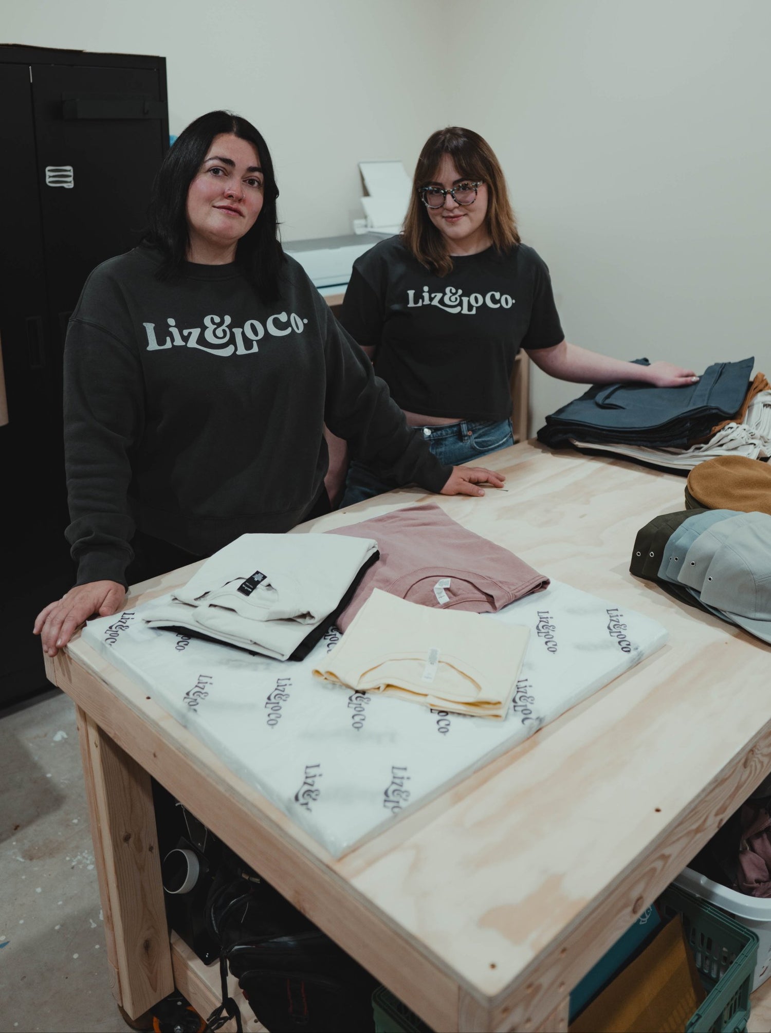 Two people wearing 'Liz & Co' shirts standing behind a table with various items on it.